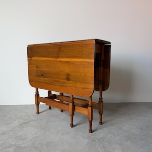 May include: A wooden drop-leaf table with a warm, medium-brown finish. The table has a rectangular top with rounded edges and is supported by four turned legs. When open, the table is approximately 30 inches long.