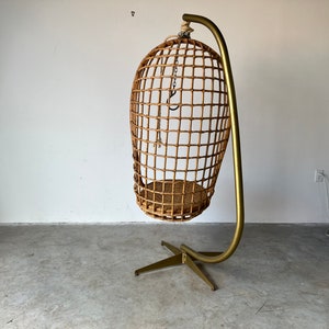 May include: A woven wicker hanging chair with a gold metal stand. The chair is suspended from the stand by a chain.