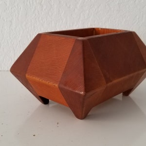 May include: A geometric wooden planter with a reddish-brown finish. The planter has a square base and a unique, faceted design.