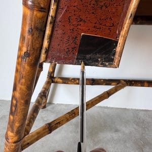 May include: Close-up of a vintage bamboo table with a red and black tabletop. The bamboo frame has a natural, aged appearance with visible knots and variations in color. The tabletop shows signs of wear.