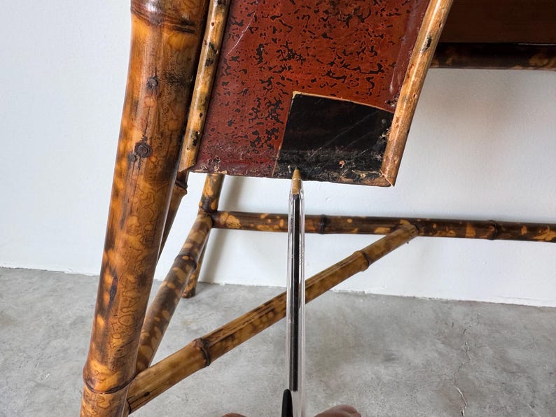 May include: Close-up of a vintage bamboo table with a red and black tabletop. The bamboo frame has a natural, aged appearance with visible knots and variations in color. The tabletop shows signs of wear.