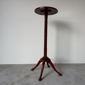 May include: A dark brown, antique-style plant stand. The stand features a round top, a long, ribbed central column, and three curved legs with claw feet. The stand is made of wood and stands approximately 30 inches tall.