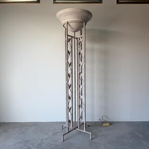May include: A tall, cream-colored floor lamp with a woven shade and a decorative metal frame. The lamp has a tiered shade and a geometric design on the support structure. The base is a simple, four-legged design.