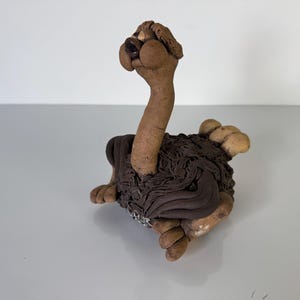 May include: A whimsical ceramic sculpture of an ostrich. The ostrich has a long, slender neck and a detailed head. The body is a textured brown, and the feet are a lighter tan color. The sculpture is on a white surface.