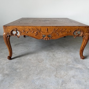 May include: A square wooden coffee table with a medium brown finish. The apron features intricate carved floral and urn motifs. The table stands on four curved cabriole legs with scroll details at the top and feet. The top surface has a flat panel.