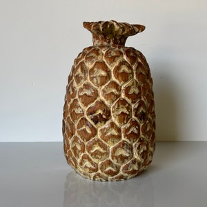 May include: A decorative pineapple sculpture with a textured, brown body and a cream-colored, carved pattern. The top features a detailed, textured crown. The sculpture is a unique home decor item.