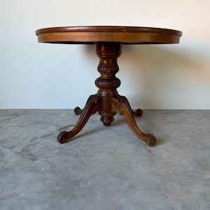 May include: A round, antique wooden table with a dark brown finish. The table has a pedestal base with carved details and three curved legs. The tabletop is approximately 30 inches in diameter.
