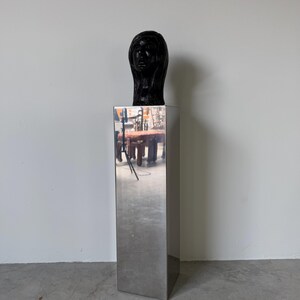 May include: A black sculpture of a woman's head atop a tall, rectangular, mirrored pedestal. The sculpture has smooth features and long hair. The pedestal reflects the surrounding environment.