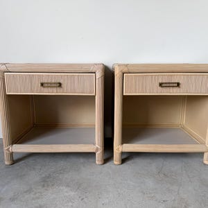 May include: A pair of light beige rattan nightstands, each featuring a single drawer with a rectangular brass-toned pull handle. The furniture pieces have a reeded texture on the frame and include an open storage shelf at the bottom.