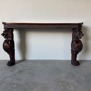 May include: A dark brown wooden console table with intricately carved lion head legs. The table has a rectangular top with a decorative edge. The lion legs are detailed with fur and clawed feet, adding a touch of elegance.