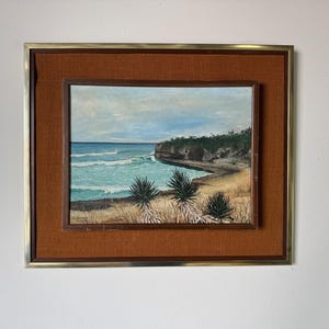 May include: An oil painting of a coastal landscape, framed in a gold-toned metal frame with a brown fabric matting. The artwork depicts a turquoise ocean with white waves, a sandy beach, and green foliage. The sky is a pale blue.