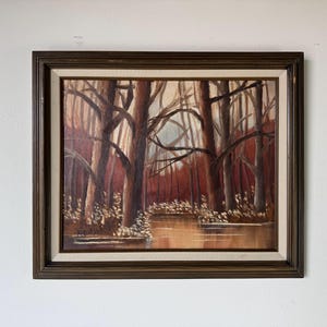 May include: An oil painting of a forest scene with tall, bare trees and a body of water. The artwork features a dark brown wooden frame and a neutral-toned mat. The color palette includes shades of brown, red, and beige.