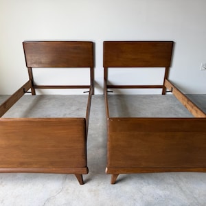 May include: A pair of matching twin-size bed frames constructed from medium-toned brown wood. Each frame features a rectangular headboard, a solid footboard, and four tapered legs. The side rails connect the headboard and footboard sections.