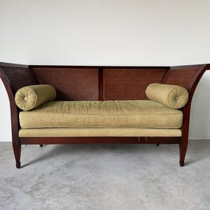 May include: A vintage loveseat with a dark wood frame, woven cane back, and olive green cushions. The loveseat features two bolster pillows and curved armrests. The design is a classic style, suitable for a living room or sitting area.