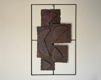 May include: A metal wall art piece featuring a geometric design of watermelon slices. The slices are cut from metal and are arranged in a modern, abstract pattern. The piece is painted in a rusty brown color and is framed in a black metal frame.