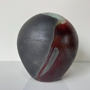 May include: A handcrafted ceramic vase with a unique design. The vase features a dark gray section on the left side, transitioning to a deep burgundy on the right. A light gray accent runs down the center. The top has a touch of green.
