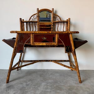 May include: Antique bamboo desk with a mirror, drawers, and side compartments. The desk features a dark red and gold color scheme. The legs are angled, and the desk has a unique, vintage design. A small mirror is centered above the drawers.