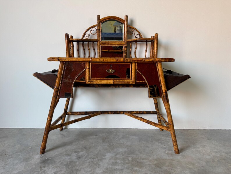 May include: Antique bamboo desk with a mirror, drawers, and side compartments. The desk features a dark red and gold color scheme. The legs are angled, and the desk has a unique, vintage design. A small mirror is centered above the drawers.