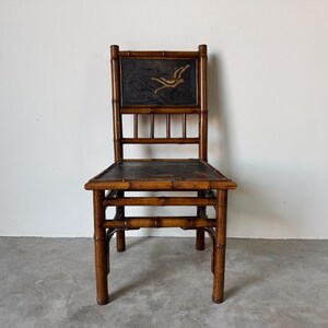 May include: A vintage bamboo chair with a dark seat and back. The back features a rectangular panel with a gold bird design on a dark background. The chair has a natural bamboo finish and a simple, elegant design.