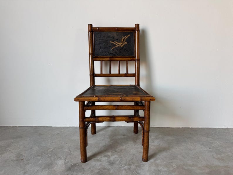 May include: A vintage bamboo chair with a dark seat and back. The back features a rectangular panel with a gold bird design on a dark background. The chair has a natural bamboo finish and a simple, elegant design.