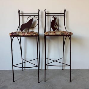 May include: Two tall, dark metal bar stools. Each stool has a floral patterned seat and a decorative cutout on the backrest. One features a howling wolf silhouette, the other a Kokopelli figure. The stools have a cross-braced design.