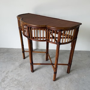 May include: A semi-circular console table constructed from brown bamboo and rattan. The piece features a woven top surface, a curved apron with vertical bamboo slats, and four slender legs connected by a lower support structure for stability.