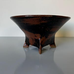 May include: A handcrafted, three-legged ceramic bowl with a flared rim. The bowl features a dark brown and black mottled glaze, creating a rustic and earthy aesthetic. This decorative piece is suitable for various uses.