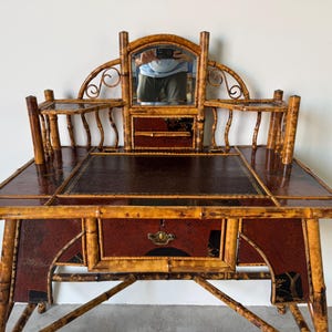 May include: Antique bamboo desk with a dark brown leather writing surface and a small mirror. The desk has a red and black painted design on the front and sides, with a drawer and decorative hardware. The desk is supported by bamboo legs.