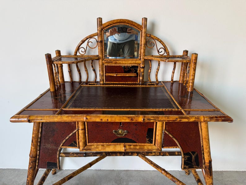 May include: Antique bamboo desk with a dark brown leather writing surface and a small mirror. The desk has a red and black painted design on the front and sides, with a drawer and decorative hardware. The desk is supported by bamboo legs.