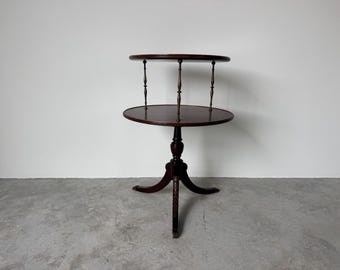 May include: A dark brown, two-tiered side table with a round top and a lower, larger round shelf. The table has three legs and is supported by three slender posts. The table is approximately 28 inches tall.