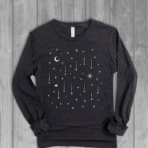 May include: Dark gray long-sleeve shirt with a white celestial design. The design features a crescent moon, stars, shooting stars, a sun, and a planet. The shirt has a crew neck and long sleeves.