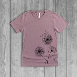 A woman's short-sleeved shirt with a dandelion design printed on the front left side seam. The shirt is a muted orchid pink mauve color, and the flower design is in black.