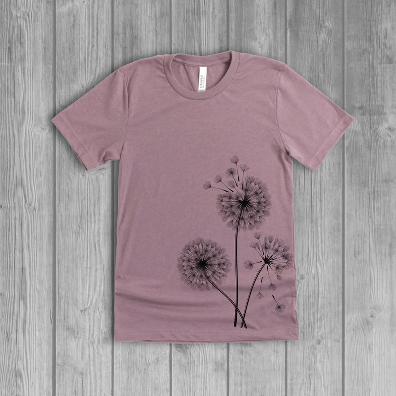 A woman's short-sleeved shirt with a dandelion design printed on the front left side seam. The shirt is a muted orchid pink mauve color, and the flower design is in black.