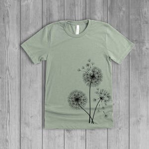 A woman's short-sleeved shirt with a dandelion design printed on the front left side seam. The shirt is a dusty sage green color, and the flower design is in black.