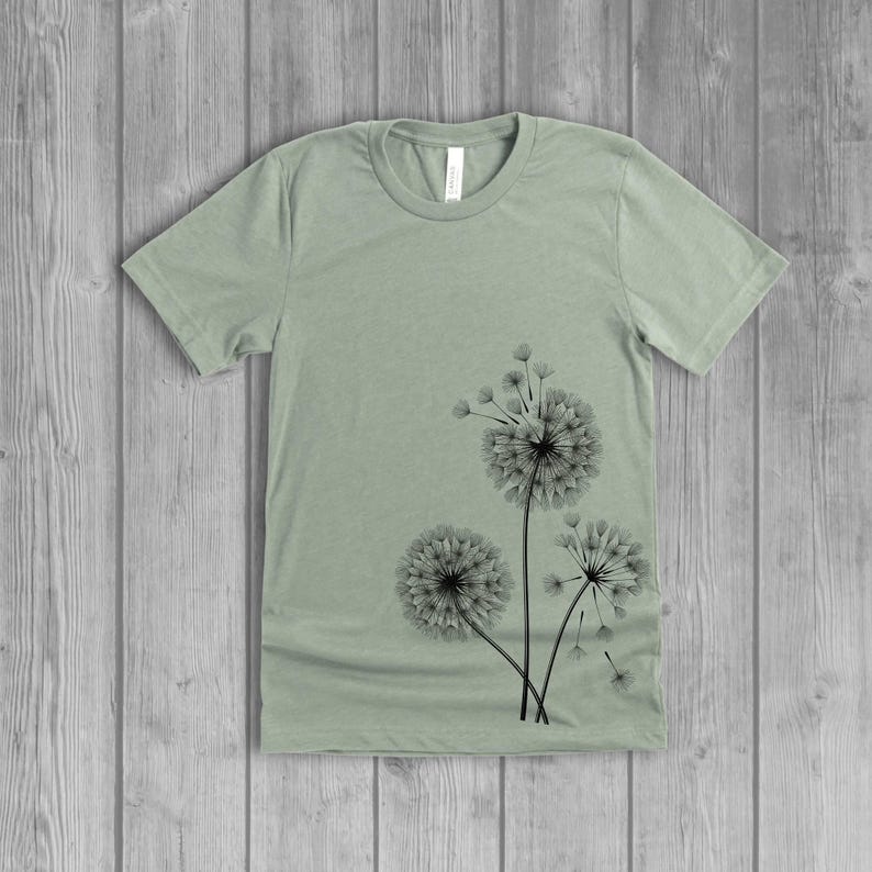 A woman's short-sleeved shirt with a dandelion design printed on the front left side seam. The shirt is a dusty sage green color, and the flower design is in black.