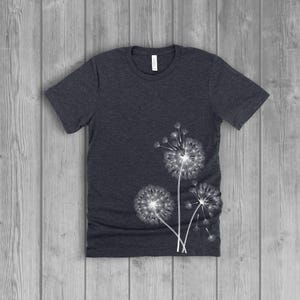A woman's short-sleeved shirt with a dandelion, with the seeds blowing in the wind, design printed on the front left side seam. The shirt is a dark charcoal grey heather color, and the flower design is in white.
