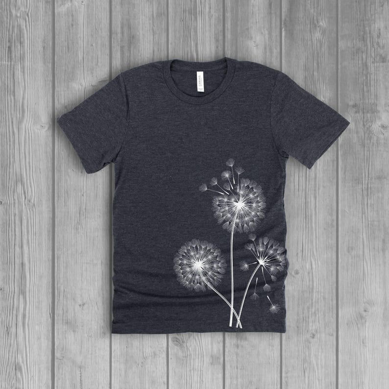 A woman's short-sleeved shirt with a dandelion, with the seeds blowing in the wind, design printed on the front left side seam. The shirt is a dark charcoal grey heather color, and the flower design is in white.