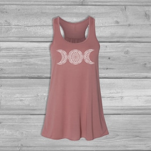 May include: A dusty rose tank top with a white triple moon mandala design. The tank top has a scoop neck and racerback straps.