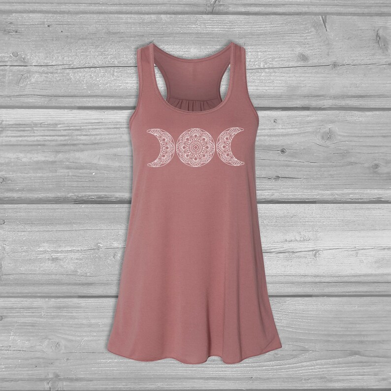 May include: A dusty rose tank top with a white triple moon mandala design. The tank top has a scoop neck and racerback straps.