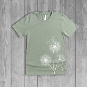 A woman's short-sleeved shirt with a dandelion design printed on the front left side seam. The shirt is a dusty sage green color, and the flower design is in white.