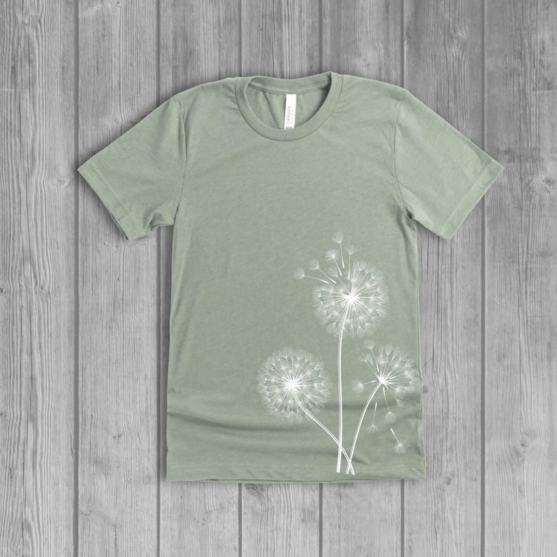 A woman's short-sleeved shirt with a dandelion design printed on the front left side seam. The shirt is a dusty sage green color, and the flower design is in white.
