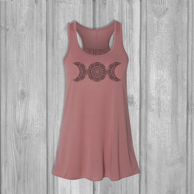 May include: A dusty rose tank top with a black triple moon mandala design. The tank top has a relaxed fit and a scoop neck.