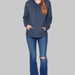 May include: A woman wearing a navy blue hooded sweatshirt with a front pocket and drawstring. She is also wearing blue jeans with a distressed knee and black and white sneakers. The background is a solid gray.