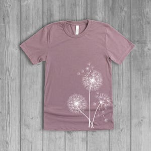 A woman's short-sleeved shirt with a dandelion design printed on the front left side seam. The shirt is a muted orchid pink mauv color, and the flower design is in white.