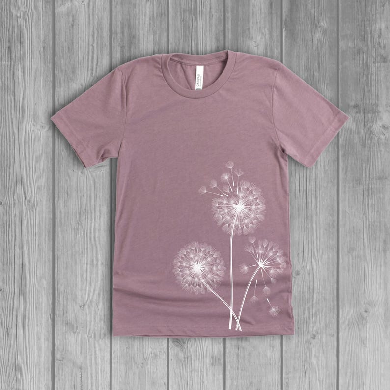 A woman's short-sleeved shirt with a dandelion design printed on the front left side seam. The shirt is a muted orchid pink mauv color, and the flower design is in white.