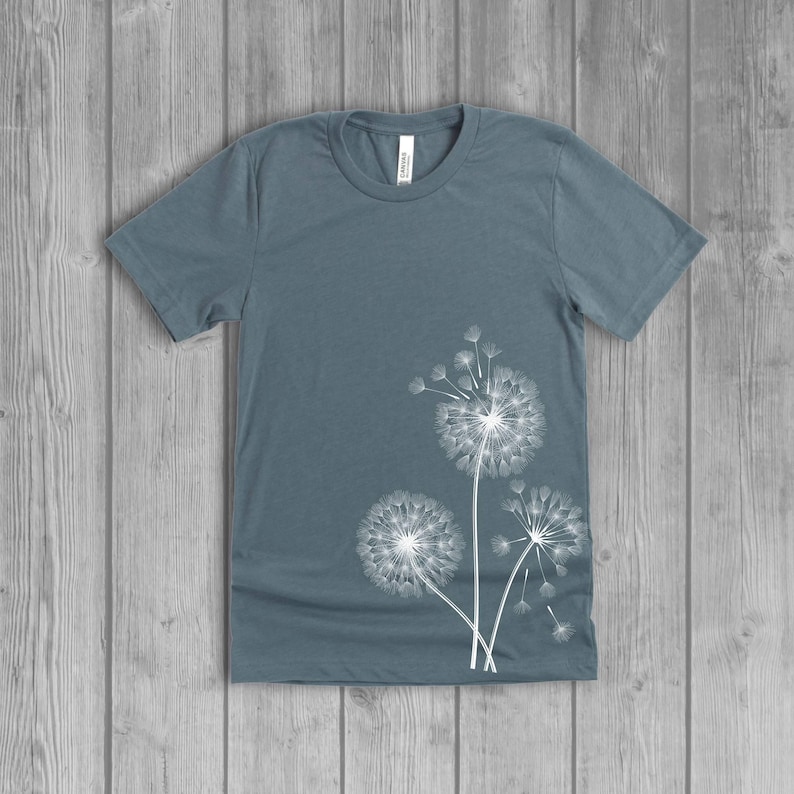 A woman's short-sleeved shirt with a dandelion design printed on the front left side seam. The shirt is a slate blue color, and the flower design is in white.