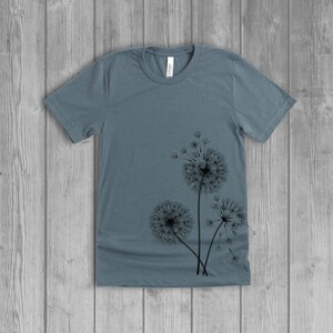 A woman's short-sleeved shirt with a dandelion design printed on the front left side seam. The shirt is a slate blue color, and the flower design is in black.
