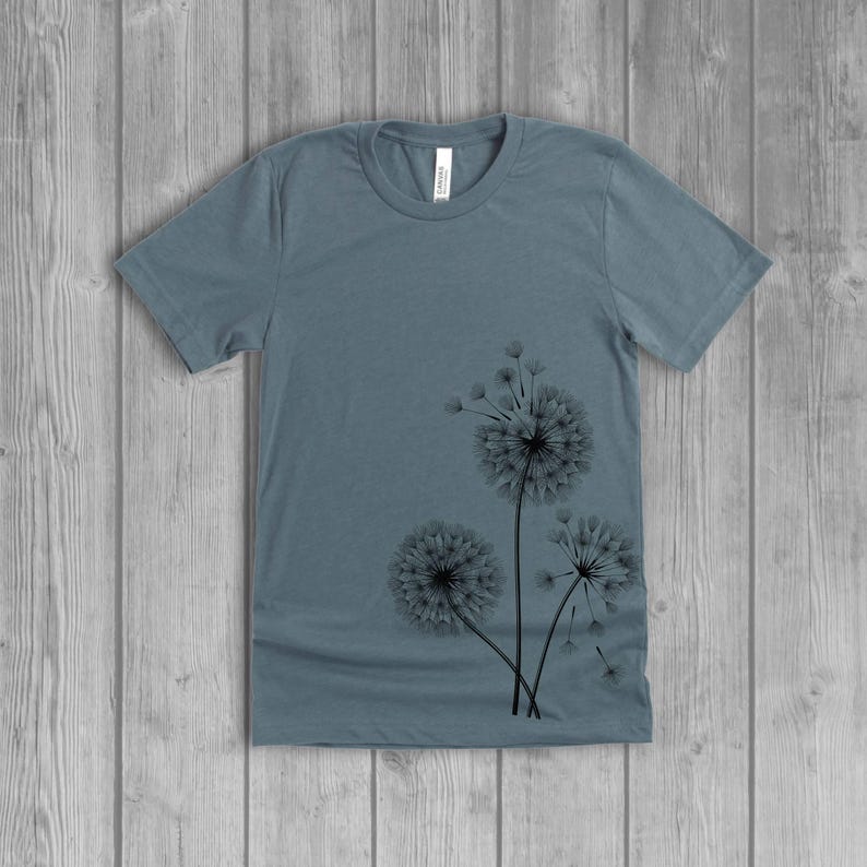A woman's short-sleeved shirt with a dandelion design printed on the front left side seam. The shirt is a slate blue color, and the flower design is in black.