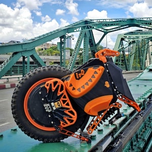 May include: A black and orange electric unicycle with a large wheel and a suspension system. The unicycle is on a green metal bridge.