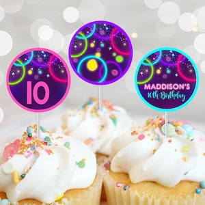 Op de afbeelding: Drie cupcake-toppers met een neon-glow-thema. De toppers hebben een paars, blauw en groen cirkelontwerp op een zwarte achtergrond met witte sterren. Eén topper heeft het nummer 10 in roze, een andere zegt "MADDISON'S 10th Birthday" in wit, en de derde heeft een kleurrijk cirkelontwerp.